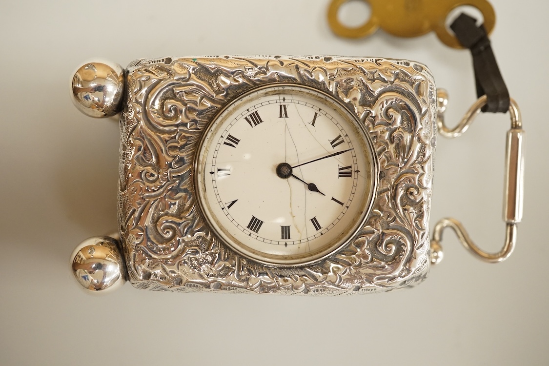 An early 20th century white metal (tests as silver) carriage timepiece, with white Roman dial (cracks) and case embossed with foliage, on four ball feet, 76mm excluding handle, with key.